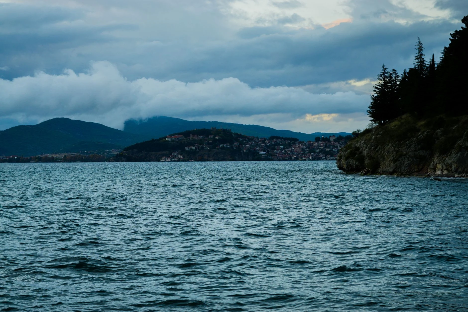 Lago Ohrid con una población y montañas bajo un cielo nublado.